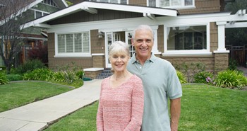 An older couple stands side by side in front of their house, sharing a moment of happiness and connection.